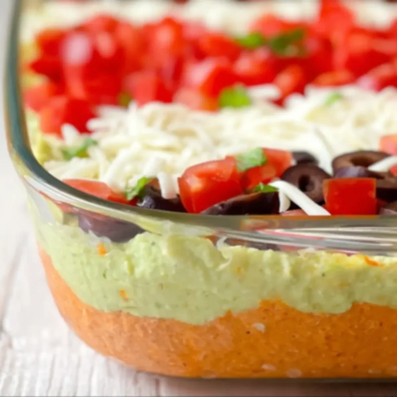 Side view of American Flag Layered Taco Dip showing distinct layers of refried beans, guacamole, sour cream, shredded cheese, tomatoes, and black olives in a glass dish.