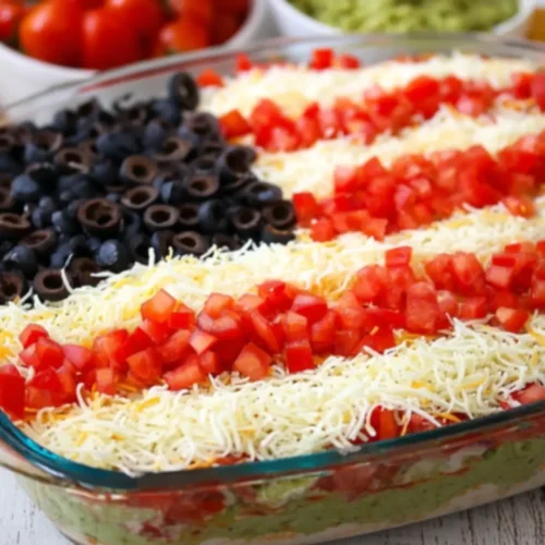 American Flag Layered Taco Dip with olives, tomatoes, and shredded cheese arranged in red, white, and blue stripes in a glass dish.