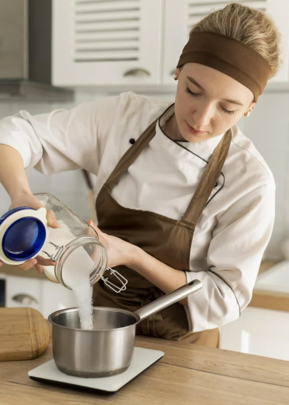 Female chef in apron pouring sugar into a saucepan while cooking in a kitchen