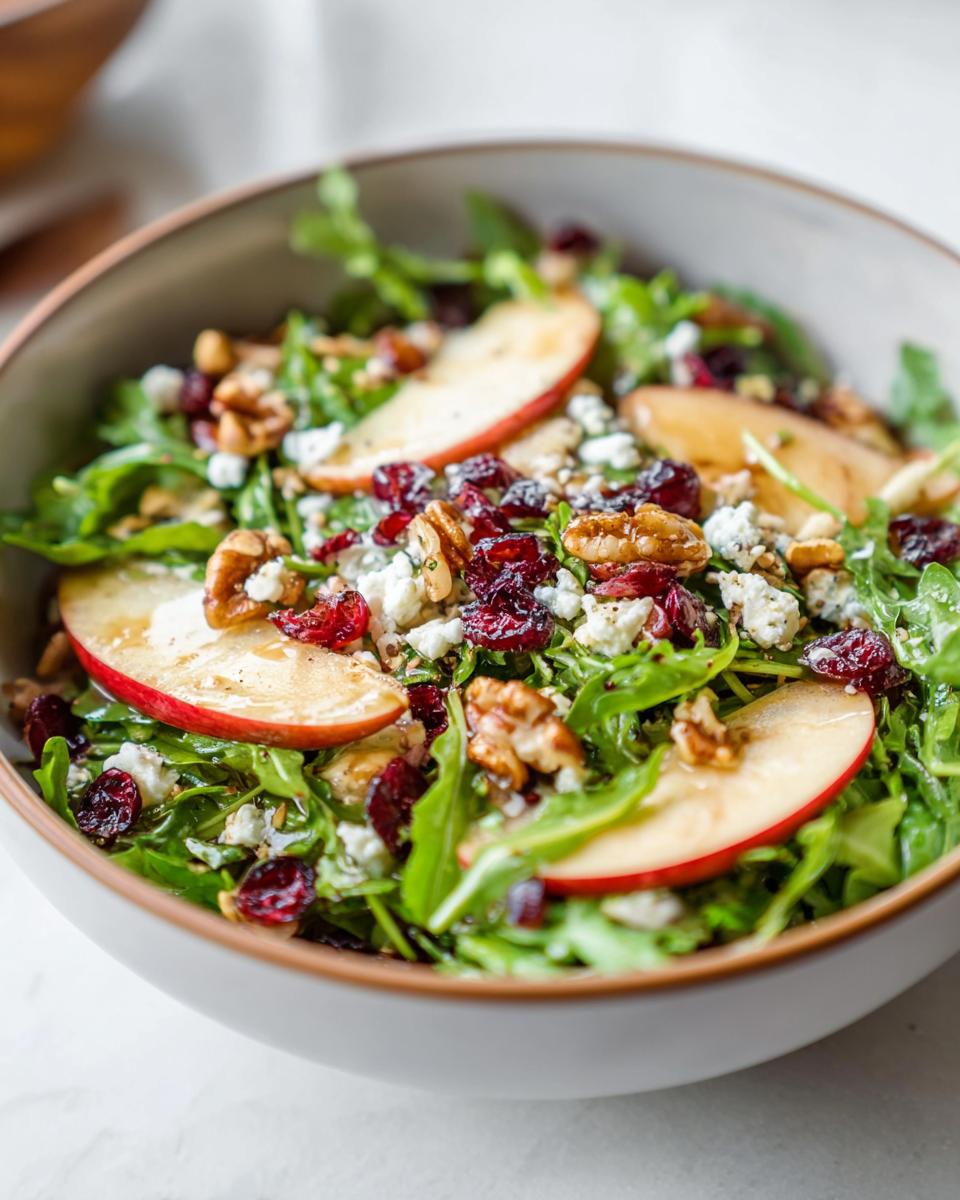 A vibrant bowl of Apple Walnut Salad featuring crisp apple slices, crunchy walnuts, dried cranberries, and crumbled blue cheese over arugula.