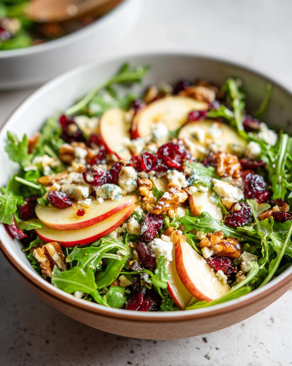 A close-up of a vibrant Apple Walnut Salad in a bowl, featuring crisp arugula, sliced apples, walnuts, dried cranberries, and crumbled blue cheese.