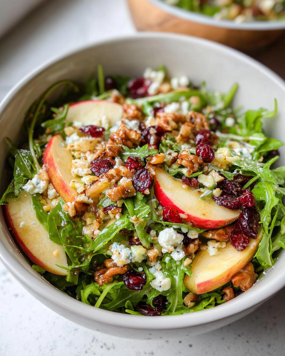A close-up of a refreshing Apple Walnut Salad in a bowl, featuring crisp apple slices, crunchy walnuts, dried cranberries, and crumbled blue cheese over arugula.