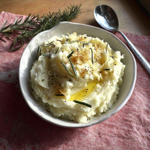 Close-up of a bowl of Best Rosemary Garlic Mashed Potatoes topped with olive oil, crispy garlic, and rosemary sprigs.