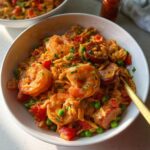 A close-up of a bowl filled with spicy Cajun orzo pasta, featuring plump shrimp, sliced sausage, peas, and diced tomatoes.