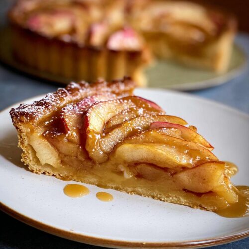 A close-up of a slice of Caramel Apple Tart, showcasing tender apple slices coated in rich caramel sauce and dusted with powdered sugar.
