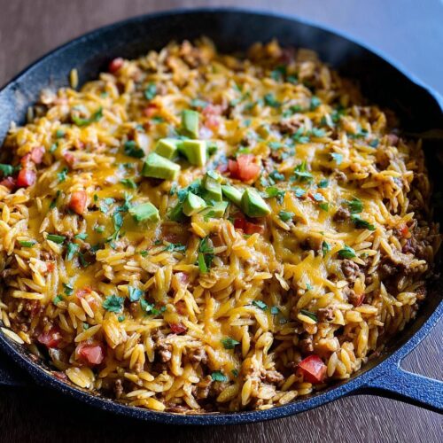 A close-up of a Cheesy Taco Orzo Skillet Dinner in a cast iron pan, topped with avocado and cilantro.