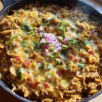 A close-up of a Cheesy Taco Orzo Skillet Dinner in a cast iron pan, topped with avocado, red onion, and cilantro.