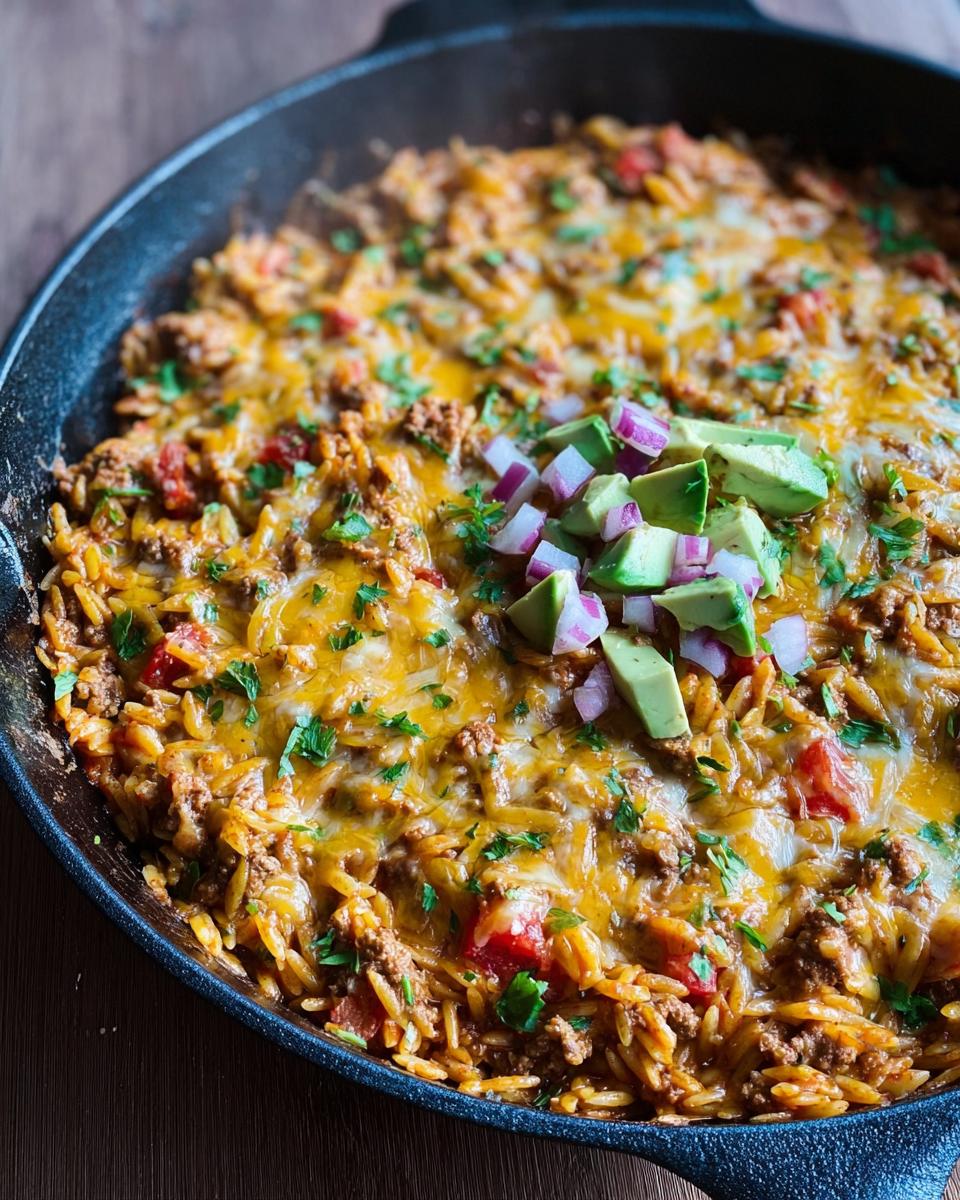 A close-up of a Cheesy Taco Orzo Skillet Dinner in a cast-iron pan, topped with melted cheese, avocado, and red onion.