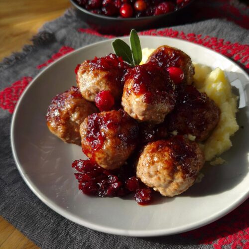 A plate of juicy Cranberry Glazed Turkey Meatballs served over mashed potatoes, garnished with cranberries and sage.