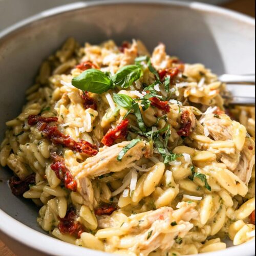A close-up of creamy pesto orzo pasta with shredded chicken, sun-dried tomatoes, and fresh basil garnish.