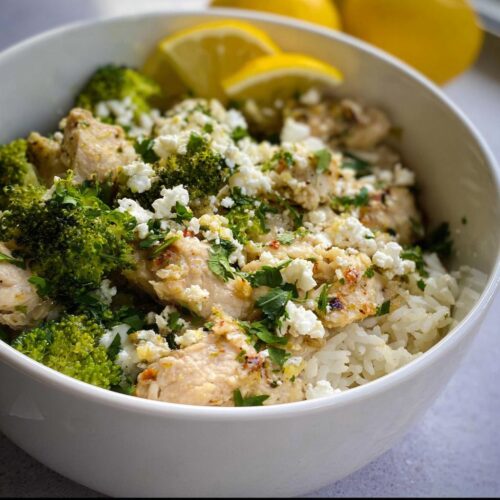 A white bowl filled with Crockpot Lemon Herb Chicken & Rice, topped with broccoli florets, crumbled feta cheese, and fresh parsley. Lemon wedges are visible in the background.
