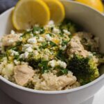 A white bowl filled with Crockpot Lemon Herb Chicken & Rice, topped with feta cheese, broccoli, and lemon slices.