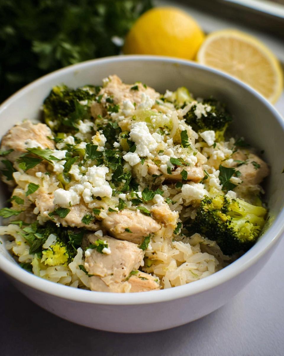 A close-up of a bowl filled with Crockpot Lemon Herb Chicken & Rice, featuring tender chicken, broccoli florets, and crumbled feta cheese.