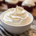 A close-up of a bowl filled with fluffy, piped Homemade Cooked Frosting, with unfrosted cupcakes in the background.