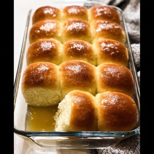 A close-up of golden brown, fluffy homemade dinner rolls baked in a glass dish with melted butter.