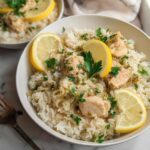A close-up of a white bowl filled with fluffy rice topped with tender chicken pieces, garnished with lemon slices and fresh parsley.