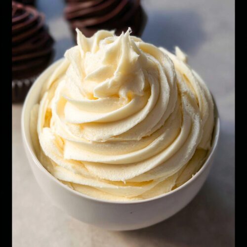 A bowl filled with smooth, swirled No-Fridge Buttercream Frosting, with chocolate cupcakes blurred in the background.