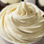 Close-up of a swirl of creamy No-Fridge Buttercream Frosting in a white bowl, with a hint of a dark cupcake in the background.