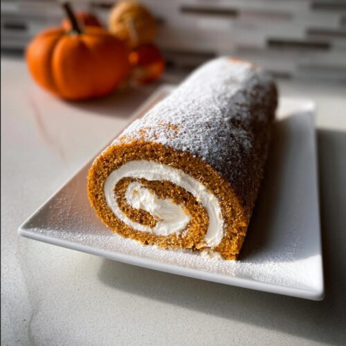 A slice of Pumpkin Roll with Cream Cheese Frosting dusted with powdered sugar on a white plate.