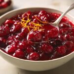 A close-up of a bowl filled with vibrant Simple Cranberry Sauce, garnished with orange zest, with a spoon lifting some.