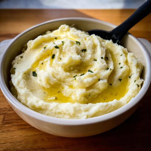 A bowl of creamy slow cooker mashed potatoes topped with melted butter and chopped parsley, with a black spoon.