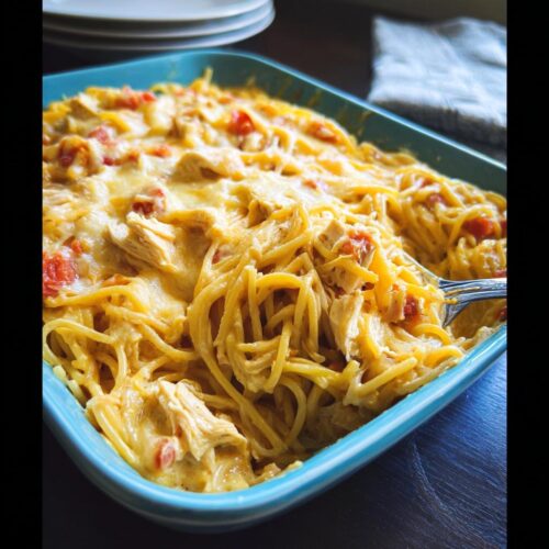 A close-up of a blue baking dish filled with cheesy Spicy Cheddar & Rotel Chicken Spaghetti Bake, with a fork lifting a portion.