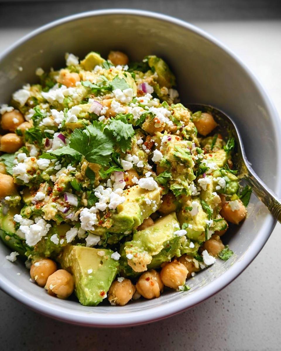 A close-up of a vibrant Avocado Chickpea Bowl topped with crumbled feta cheese, cilantro, and red onion.