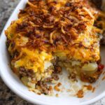 A serving dish showing a slice taken out of a Cheesy Hobo Casserole topped with melted cheddar and crispy fried onions.
