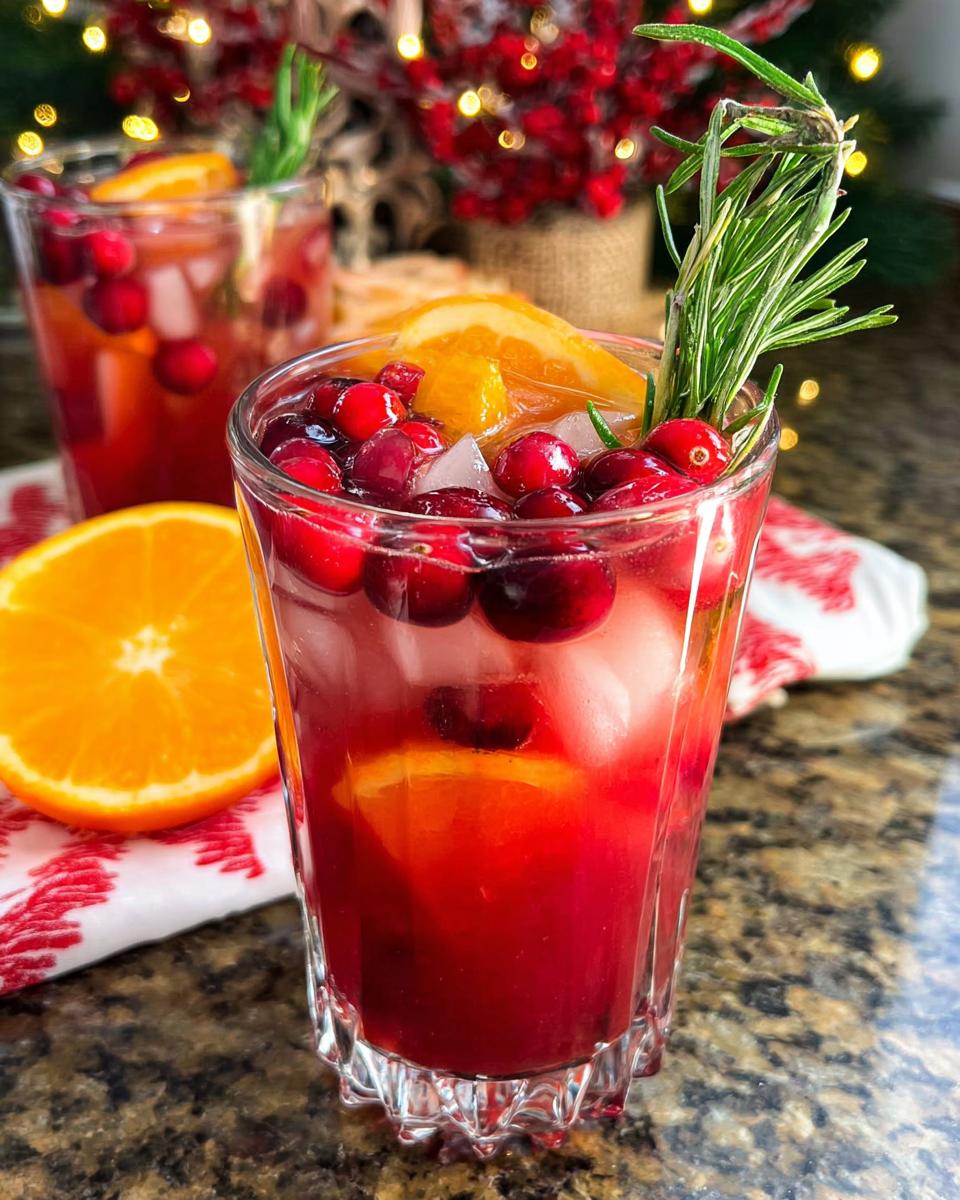 Close-up of a glass filled with Christmas Punch Mocktail, ice, fresh cranberries, orange slices, and a rosemary garnish.