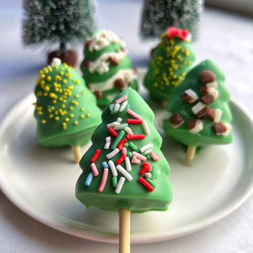 A close-up of green Christmas Tree Cake Pops decorated with festive sprinkles on a white plate.