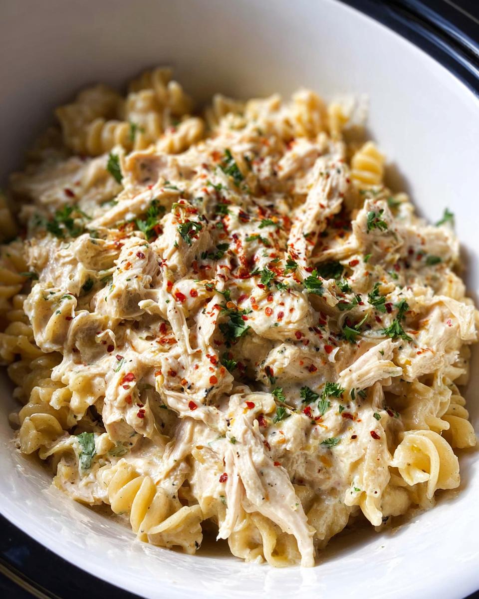 Close-up of creamy Crockpot Garlic Parmesan Chicken Pasta with fusilli noodles and shredded chicken, topped with parsley and red pepper flakes.