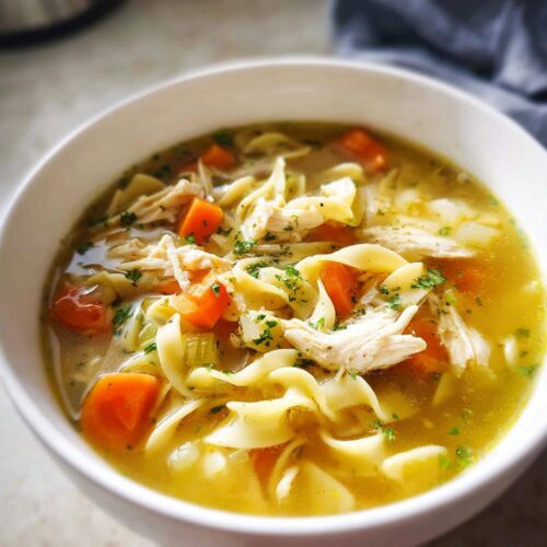 Close-up of a white bowl filled with Easy Chicken Noodle Soup featuring shredded chicken, carrots, and wide egg noodles.
