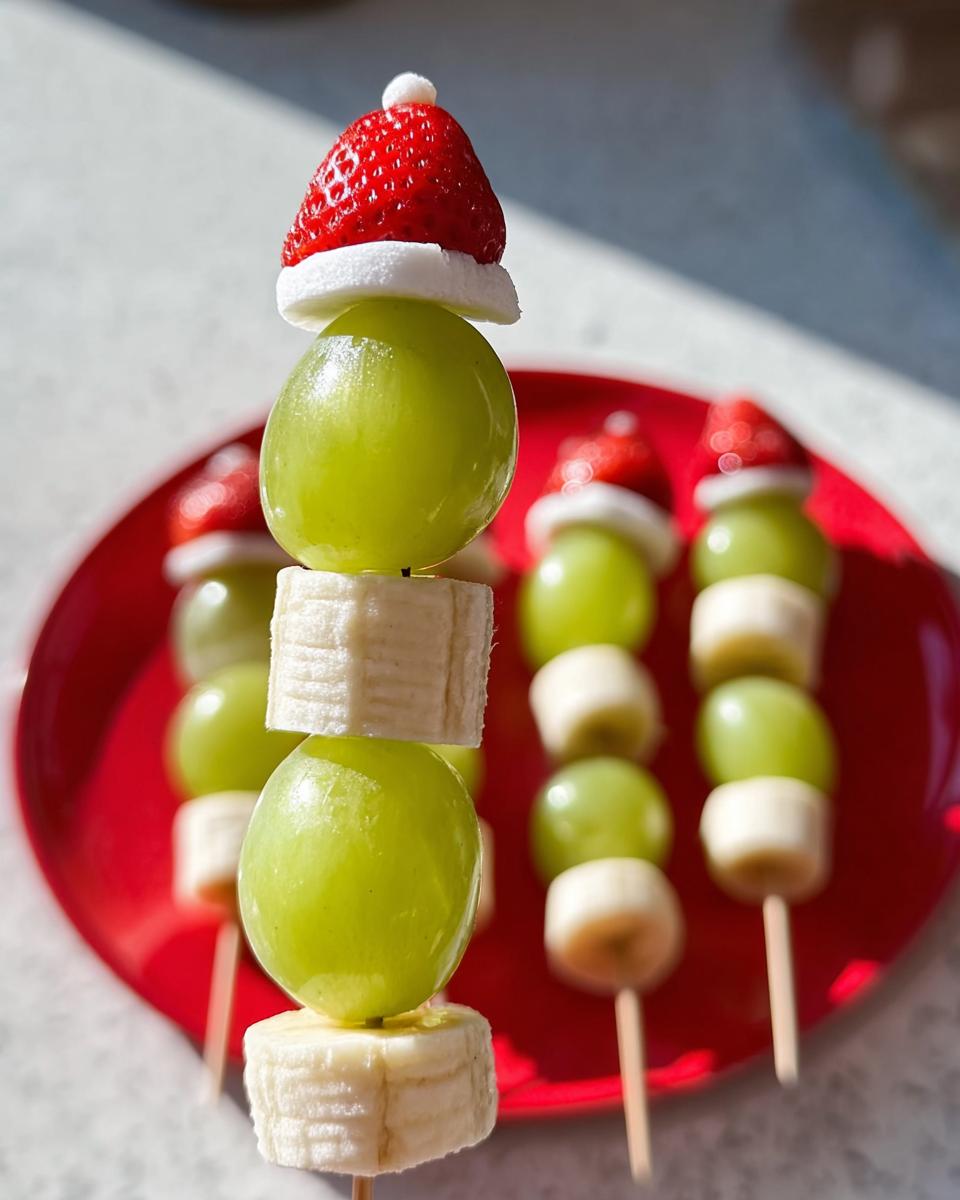 Close-up of Grinch Fruit Kebobs featuring green grapes, banana slices, and strawberries topped with marshmallow 'Santa hats'.