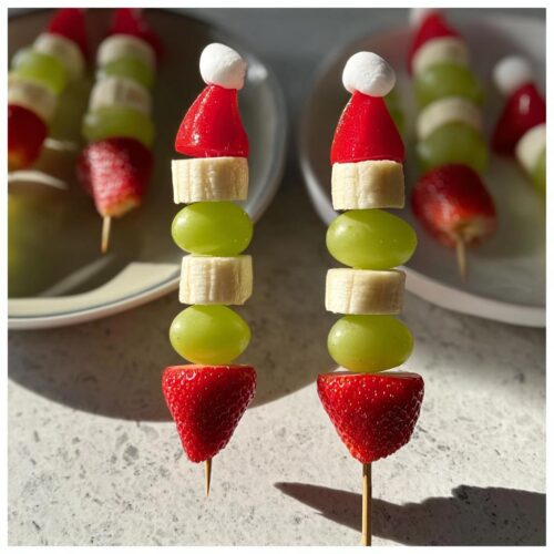 Close-up of two Grinch Fruit Kebobs topped with Santa hats, made of strawberries, grapes, and bananas.