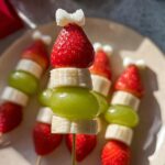 Close-up of festive Grinch Fruit Kebobs featuring strawberries, banana slices, and green grapes, topped with a white bow.