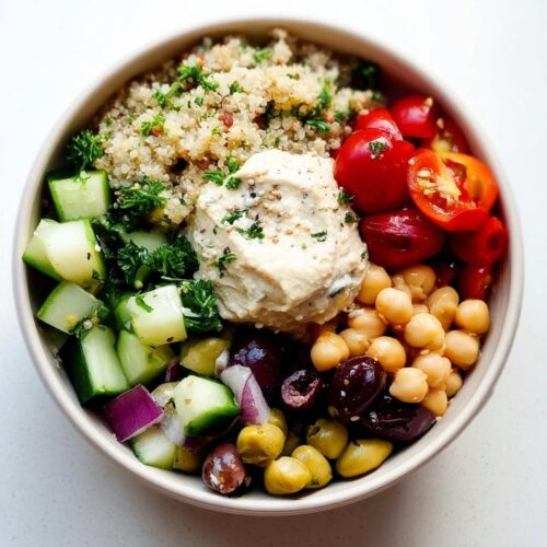 Overhead view of a bowl containing ingredients for a Healthy Mediterranean Snack Box: quinoa, hummus, chickpeas, olives, tomatoes, and cucumber.