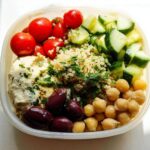 Overhead view of a clear container holding components for a Healthy Mediterranean Snack Box: hummus, cherry tomatoes, cucumber, quinoa, olives, and chickpeas.