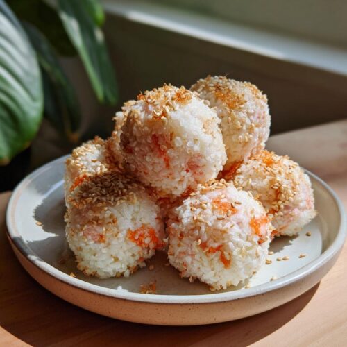 A stack of six Healthy Sushi Lunch Balls coated in toasted sesame seeds on a light ceramic plate.