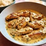 Close-up of golden-brown chicken tenders smothered in a creamy, garlic sauce, served in a white bowl next to rice.