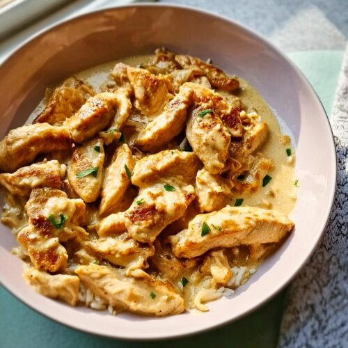 Close-up of sliced, seared chicken pieces in a creamy garlic sauce served over white rice, garnished with parsley.