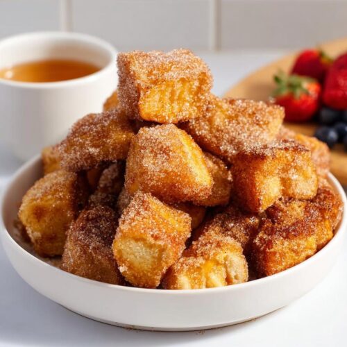 A white bowl overflowing with golden brown Air Fryer Churro French Toast Bites coated in cinnamon sugar.