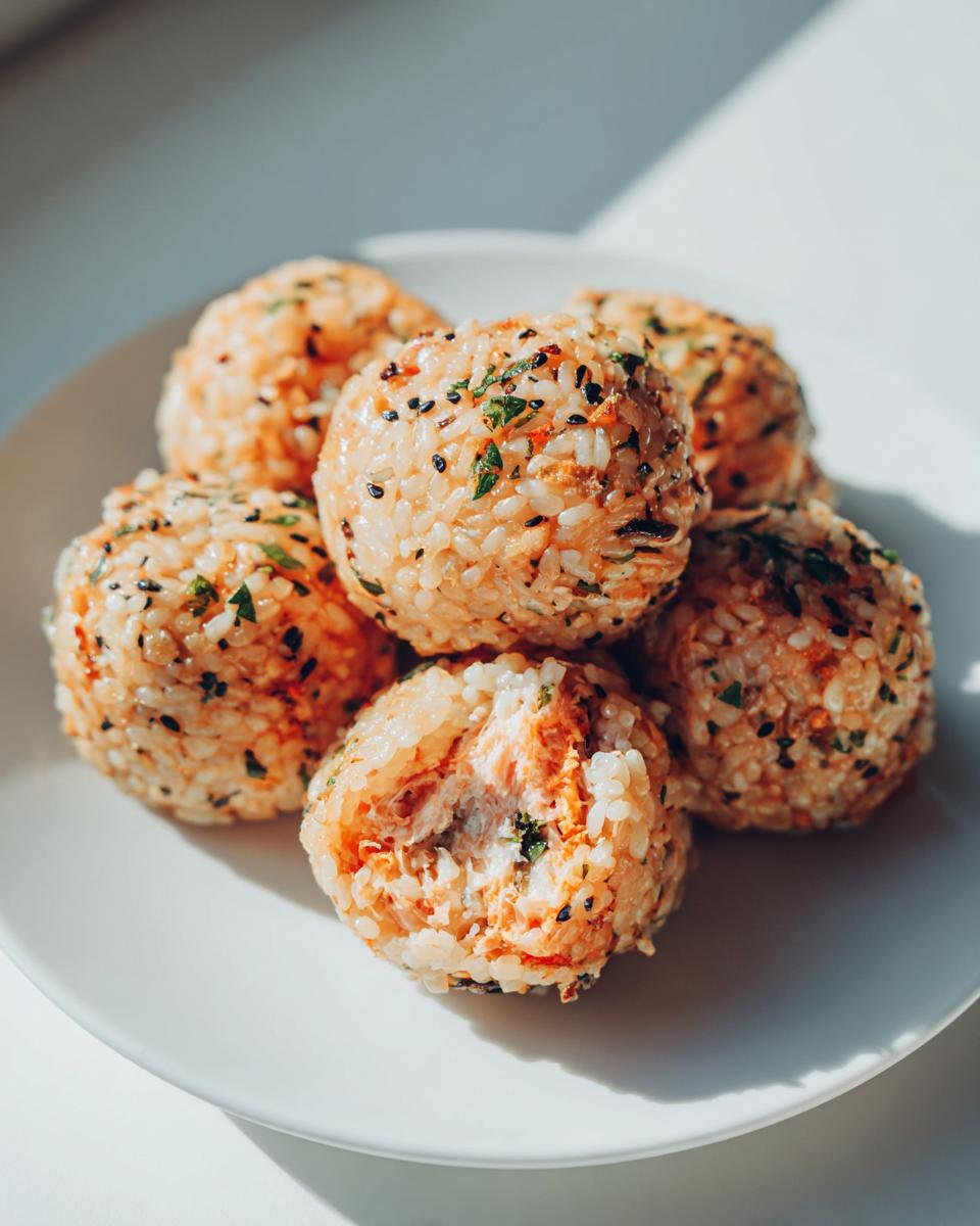 A stack of six seasoned Bento Box Sushi Balls on a white plate, one is broken open showing a salmon filling.