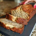 A loaf of Best Ever Sour Cream Banana Bread, sliced on a dark board, with a hand reaching toward it.
