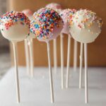 Several finished Birthday Cake Pops dipped in white and pink coating and covered in rainbow sprinkles, standing upright.