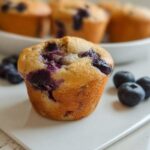 A single, golden-brown Blueberry Cottage Cheese Muffin sits on a white plate with fresh blueberries scattered nearby.