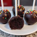 Close-up of three Brownie Batter Cake Pops on a white plate, one cut open revealing the dense, dark interior.