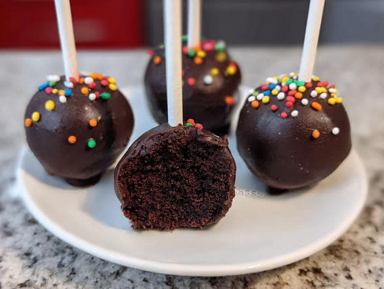 Close-up of three Brownie Batter Cake Pops on a white plate, one cut open revealing the dense, dark interior.