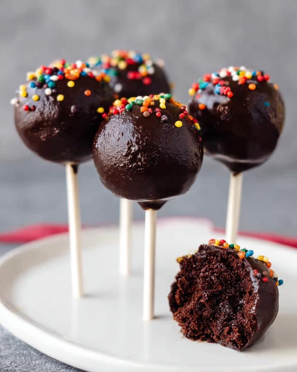 Four rich, chocolate-dipped Brownie Batter Cake Pops topped with colorful sprinkles, one is bitten into.