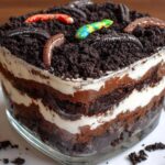 Close-up of a layered Brownie Dirt Cake in a glass dish, topped with crushed cookies and gummy worms.