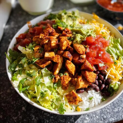 A vibrant Chicken Taco Bowl featuring seasoned chicken, rice, black beans, lettuce, salsa, guacamole, and cheese.